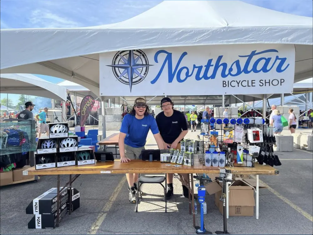 Two Northstar members posing behind a table of merchandise under a white Northstar pavilion tent